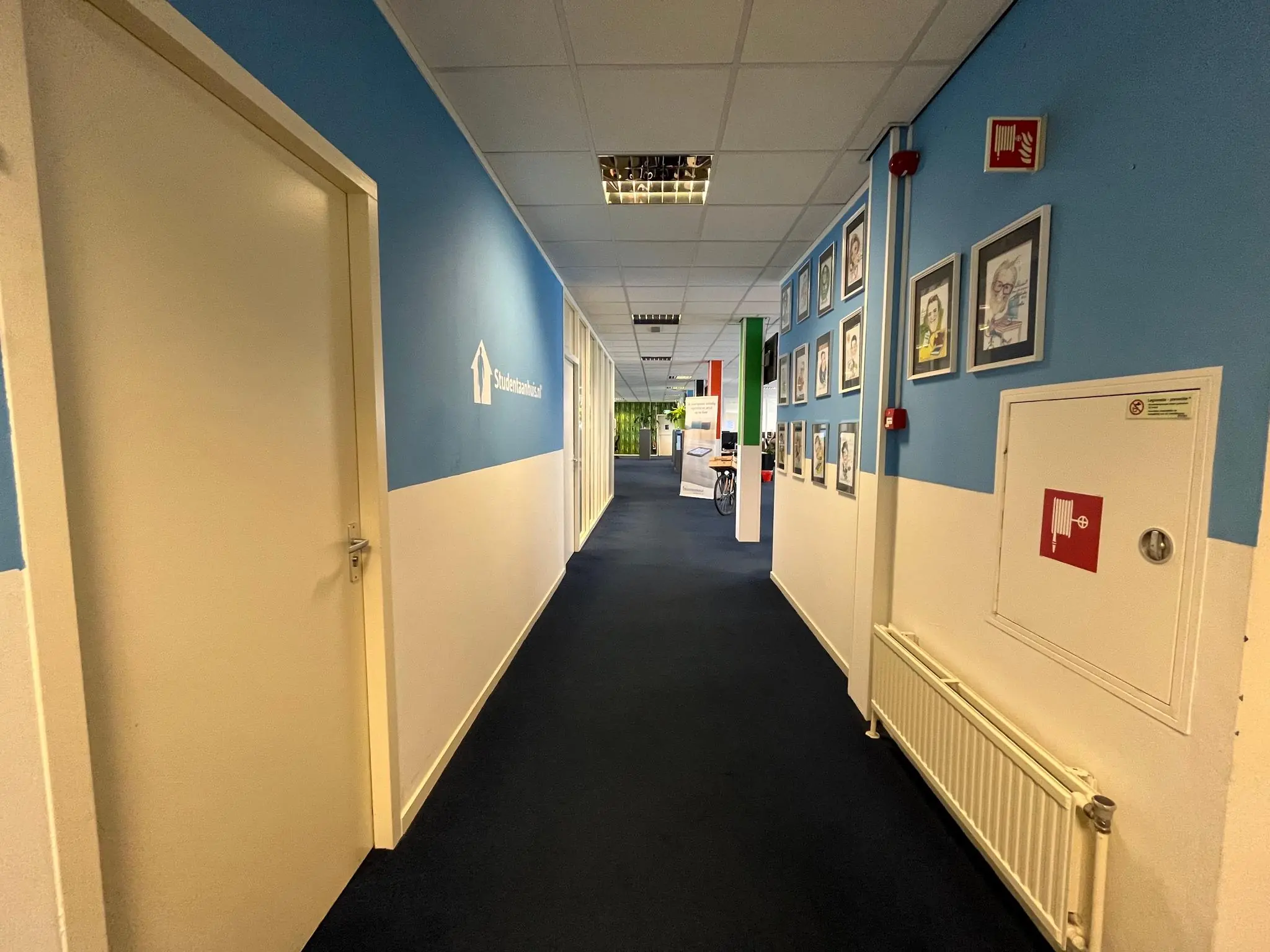 Een gang in een kantoorpand aan de Jan van Eycklaan met blauwe muren, portretfoto’s aan de wand en het logo van Studentenhuisvesting.